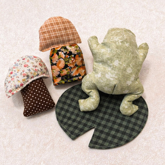 Two fabric mushroom toys, One fabric frog, and one fabric lily pad with different cottage core cotton patterns on a textured surface