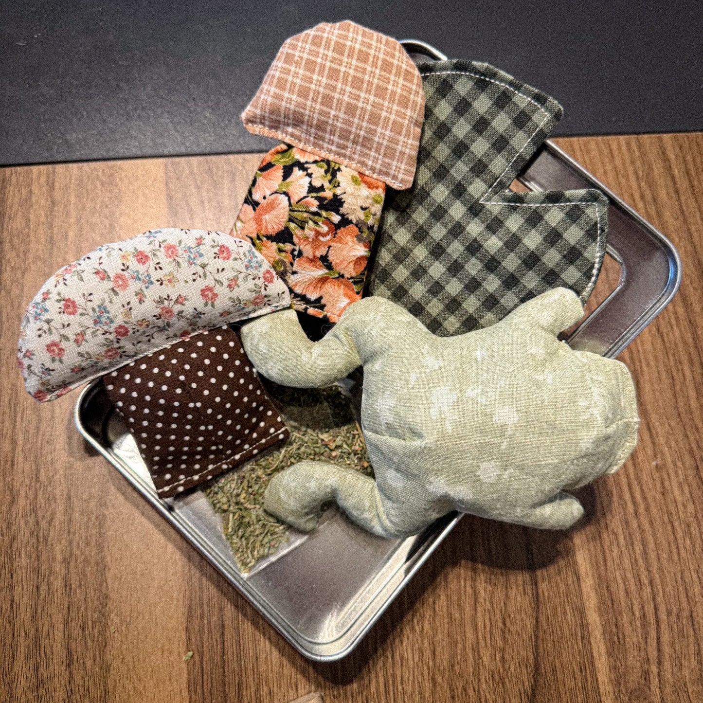 Small green frog plush cat toy with patterned mushroom toys and a lily pad in a tin on a wooden surface