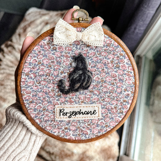 Embroidery hoop with floral pattern, small tabby cat figure, and 'Persephone' label held by a person.