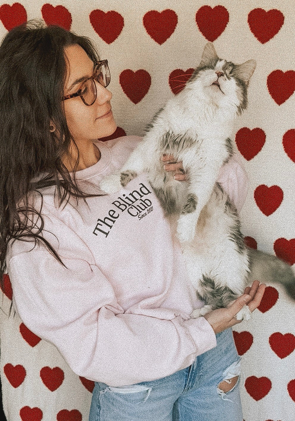 Donna (owner) holding a fluffy cat (archie) against a heart-patterned background wearing a pink "The Blind Cat Club" crewneck